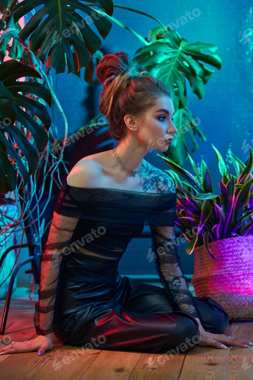 Preview: Young woman, wearing a black sheer dress, poses with tropical plants in front of a blue and red neon