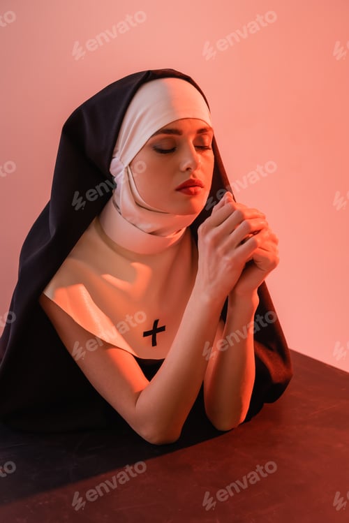 Preview: young nun praying with closed eyes on black surface isolated on pink