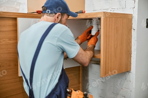 Preview: Male plumber in cap repairing pipe in kitchen