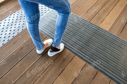 Preview: Female legs in denim jeans pants and light summer sneakers standing on wooden floow