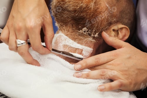 Preview: A customer sitting in the barber's chair, having a wet shave by a barber using a cut throat razor.