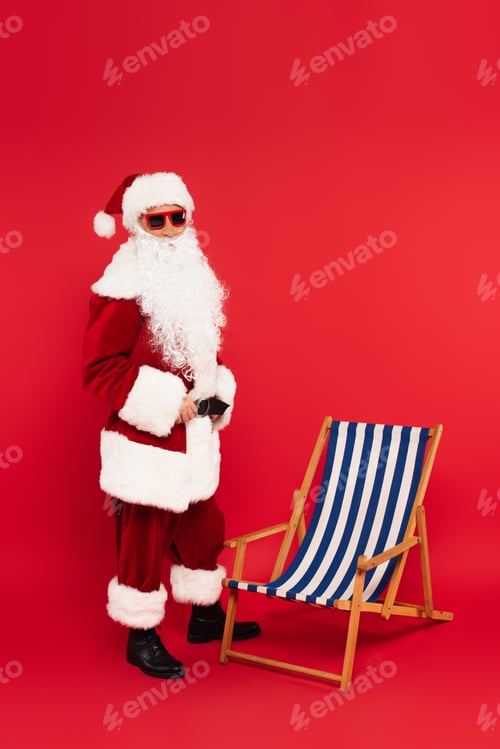 Preview: Santa claus in costume and sunglasses standing near deck chair on red background