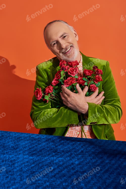 Preview: overjoyed senior male model in green velvet blazer embracing bouquet of red roses and smiling