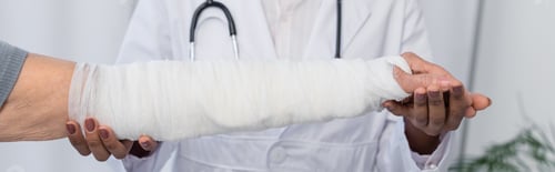 Preview: Cropped view of african american traumatologist holding hand of patient in plaster bandage, banner