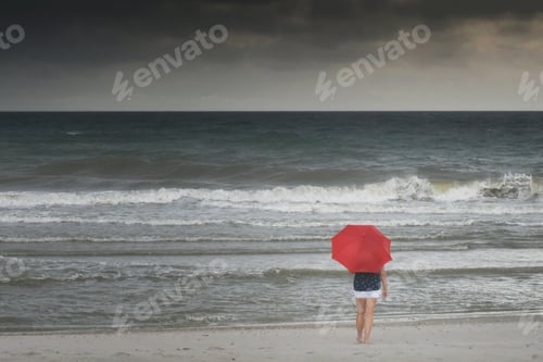Preview: A woman carrying a red umbrella walking towards stormy ocean on rainy day at the beach