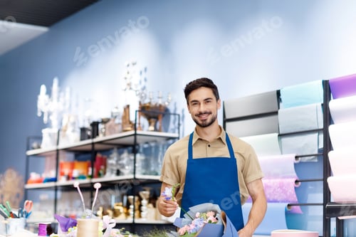 Preview: handsome bearded young florist in apron holding bouquet while working in flower shop