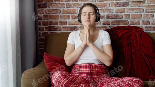 Preview: Young woman wearing headphones meditating on sofa in living room