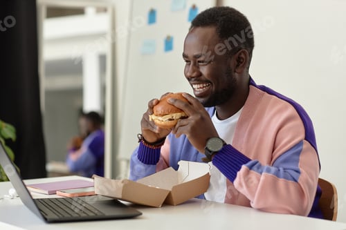 Preview: Side view smiling black man eating takeout burger at workplace in office