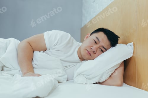 Preview: Sad Asian lying in a bed under a blanket, a depressed man in the morning