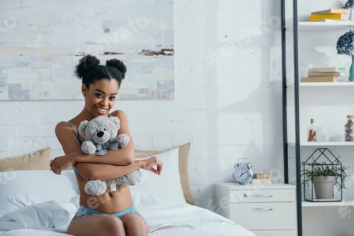 Preview: smiling african american girl hugging teddy bear while sitting on bed in morning