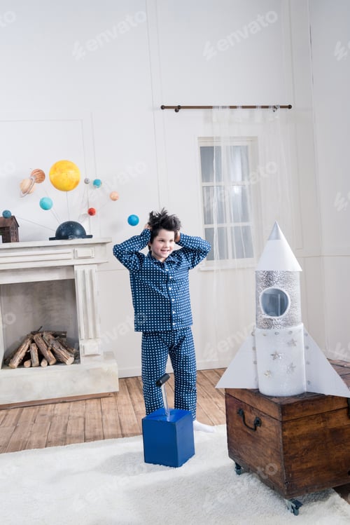 Preview: Shocked little boy in pajamas looking at toy rocket indoors