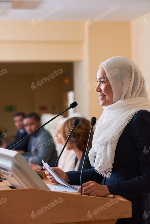 Preview: Pretty young smiling muslim female speaker standing by tribune