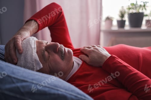 Preview: A sick senior male checking thermometer lying with wet towel on his forehead