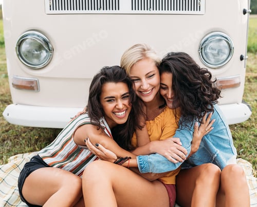 Preview: Three happy women embracing each other sitting in front of a minivan during a vacation
