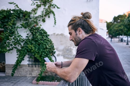 Preview: Eye-level shot of an attractive Caucasian man using his smartphone while leaning on a metal fence