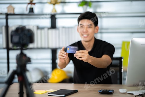 Preview: Asian male social media influencer at work place home recording vlog while showing bank card