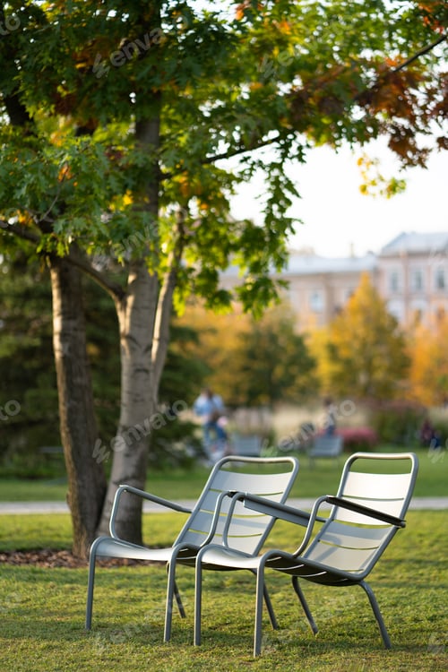 Preview: Green iron chairs on green lawn in empty public space. Autumn season.