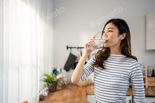 Preview: Asian young woman drinking a glass of water in kitchen at home.