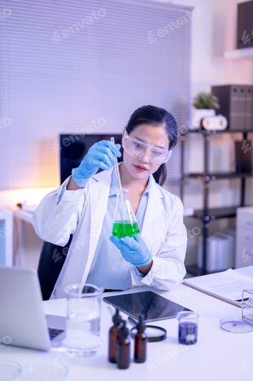Preview: Woman scientist researcher wearing goggles holding glass flask testing liquid at table in lab room.