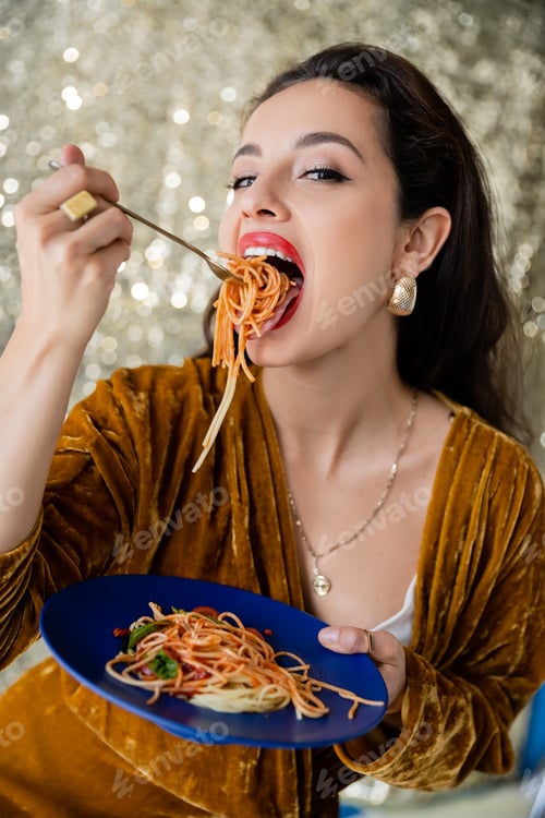 Preview: woman in velour dress eating tasty spaghetti and looking at camera on shiny silver background