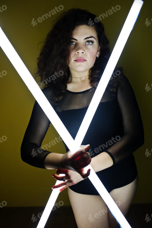 Preview: Curly brunette plus size model with two long led lamp against yellow wall.
