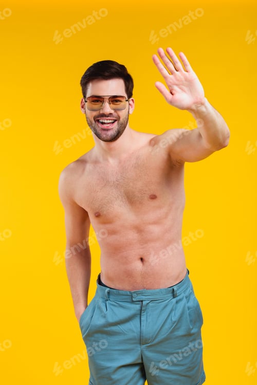 Preview: handsome shirtless young man in shorts and sunglasses waving hand and smiling at camera isolated on