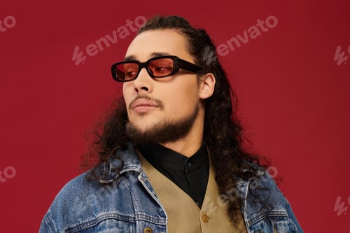 Preview: Handsome young man with a beard showcases unique style against a bold red backdrop