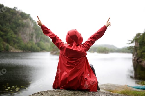 Preview: behind of person with red rain jacket raising arms and pointing up by lake