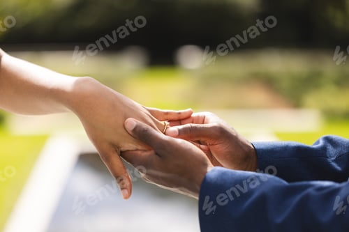 Preview: Hands of african american bride and groom placing ring on finger at wedding ceremony in sunny garden