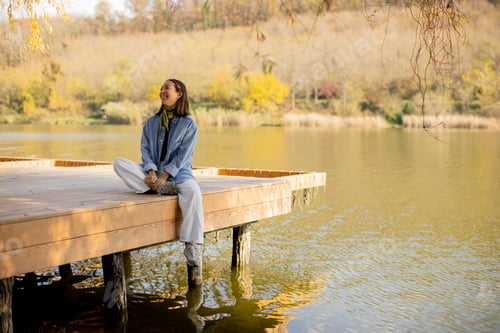 Preview: Woman enjoys serene moment by the water on a sunny autumn day in a peaceful lake setting