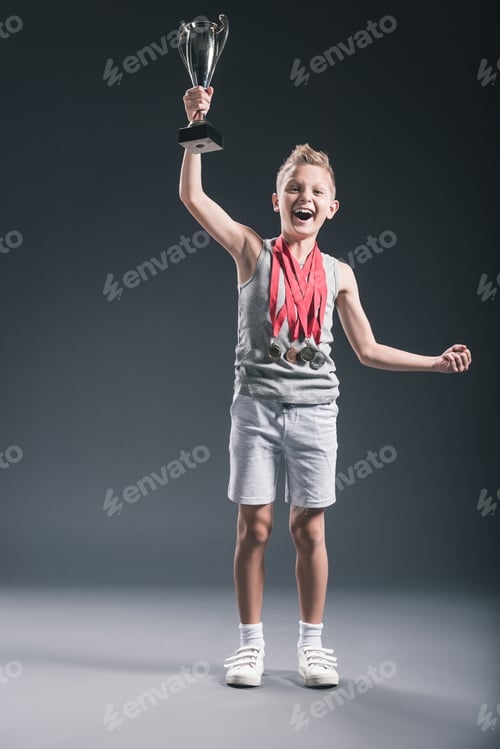 Preview: happy boy in sportswear with medals and champions cup gesturing on dark background