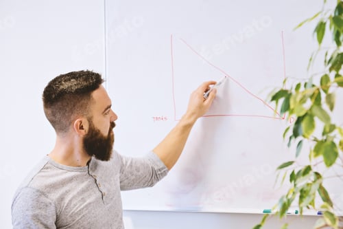 Preview: Bearded hipster, caucasian teacher or student draws chart on whiteboard during lesson in classroom