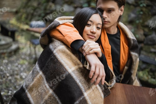 Preview: Young man hugging asian girlfriend in blanket on terrace of cafe