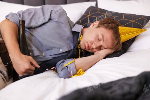 Preview: Man with bottle of alcohol suffering from hangover on bed indoors