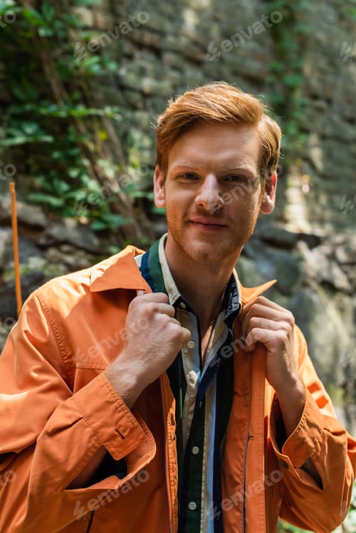 Preview: joyful redhead man in orange jacket smiling and adjusting collar on jacket outside
