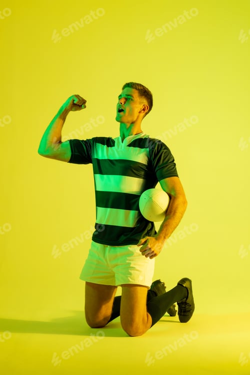 Preview: Happy caucasian male rugby player raising hands over yellow lighting