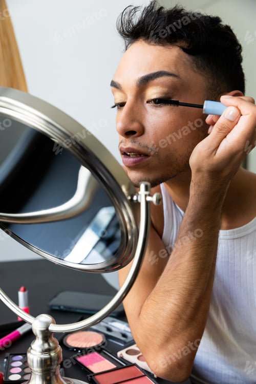 Preview: Biracial transgender man looking in mirror and putting on make-up, applying mascara