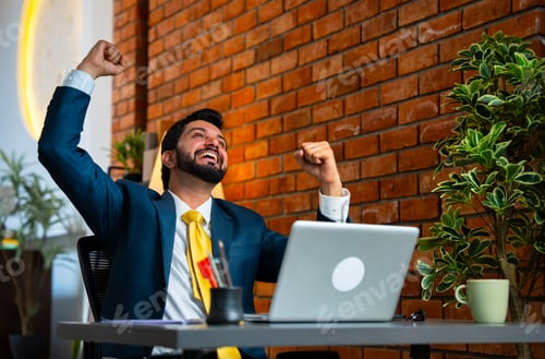 Preview: Smiling Indian businessman celebrates a big win while working on laptop in cozy office setup