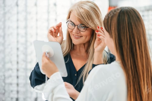 Preview: Looking at herself in the mirror. Woman choosing glasses in the store and getting help by assistant