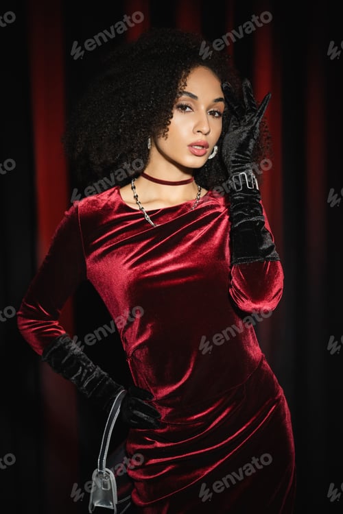 Preview: Pretty african american model posing in velvet dress and holding handbag near burgundy drapery