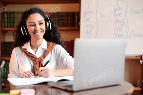 Preview: Woman sitting at desk, using laptop and writing in notebook