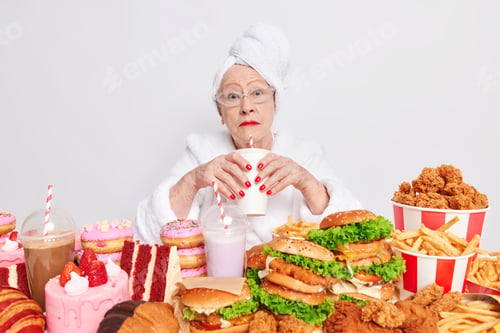 Preview: Surprised elderly lady with red lipstick has imbalancd nutrition eats different tasty junk food drin