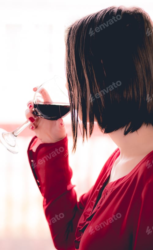Preview: young millennial woman in red shirt and red fingernail polish enjoying a glass of red wine at home