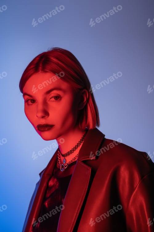 Preview: portrait of trendy woman in leather jacket and necklaces looking at camera in red light isolated