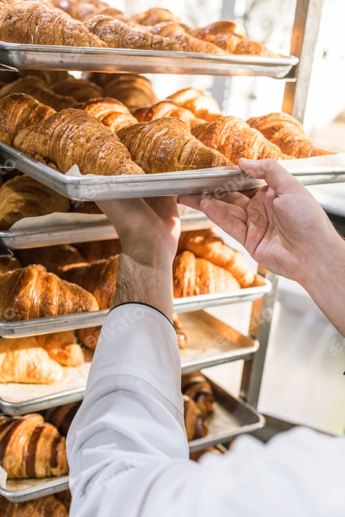 Preview: close up of male baker putting baking tray with croissants on rack