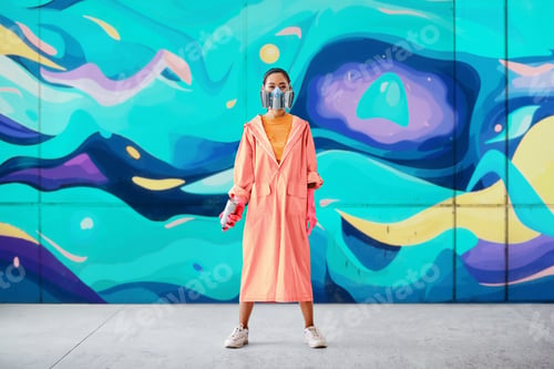 Preview: Full length portrait of female street artist in respirator mask standing near graffiti wall