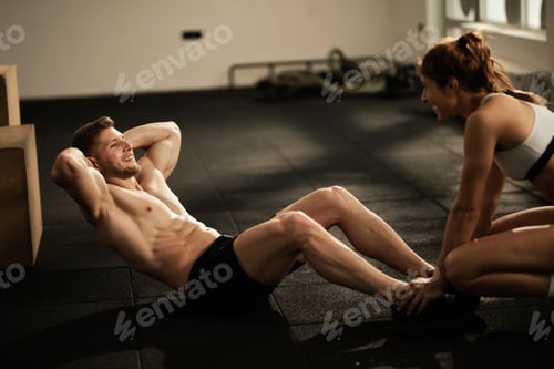 Preview: Athletic man exercising sit-ups with assistance of female coach in a gym.