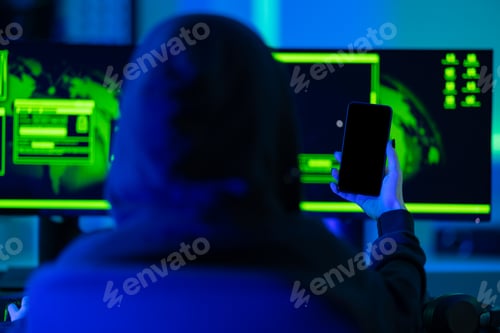 Preview: Hacker using smartphone in front of dual monitors with green glow