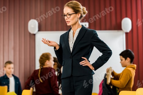 Visualização: jovem professor de óculos em pé com a mão na cintura enquanto crianças empacotam mochilas atrás