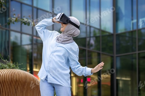 Preview: Smiling muslim woman experiencing virtual reality headset outdoors in urban setting